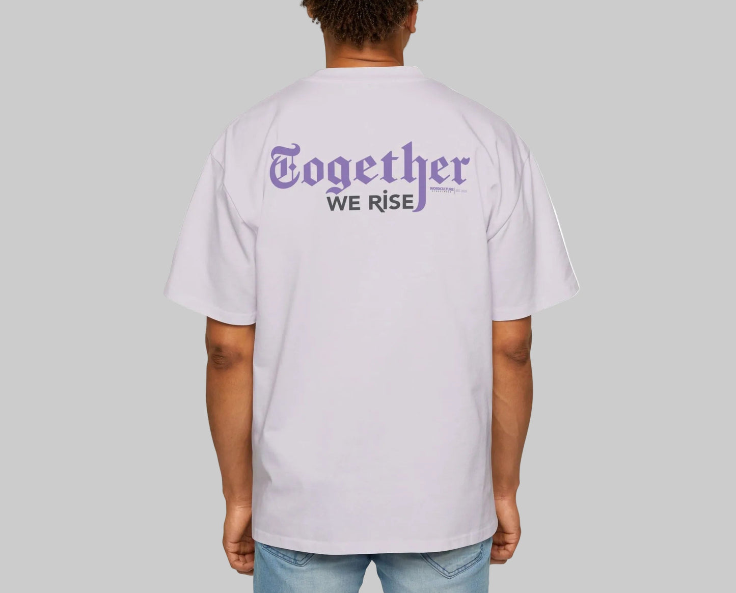 Person wearing a light purple t-shirt with 'Together We Rise' text on the back, standing against a gray background. Paired with light washed jeans.